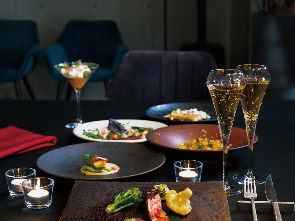 観光案内所のカウンターで、女性スタッフが地図を広げて若い男女に説明している。カウンターには、淡路島の観光やグルメに関するパンフレットや商品が並んでおり、背景には海岸線の大きな写真パネルが設置されている。