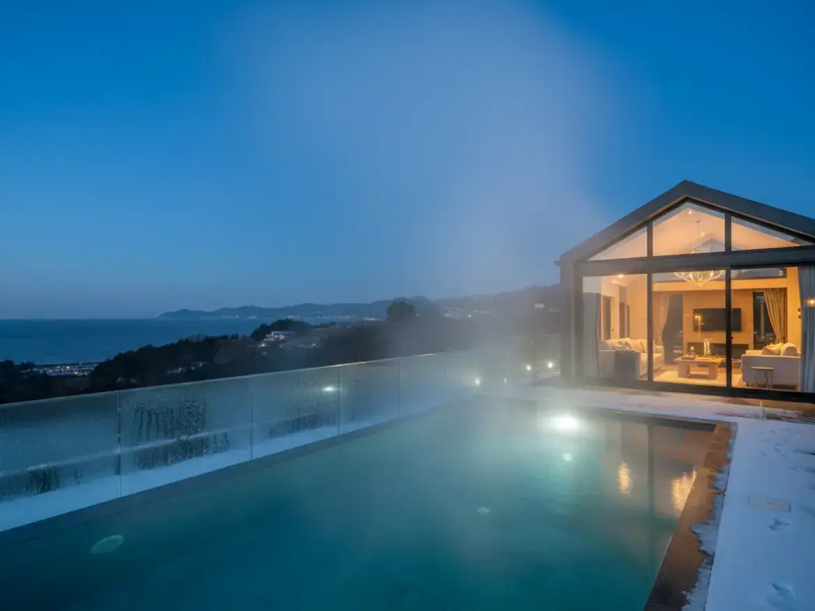 夕暮れ時、海を見下ろす家の屋外プールと、雪が積もったプールサイド。プールからは湯気が立ち上っており、大きな窓のある家の中には明かりが灯っている。