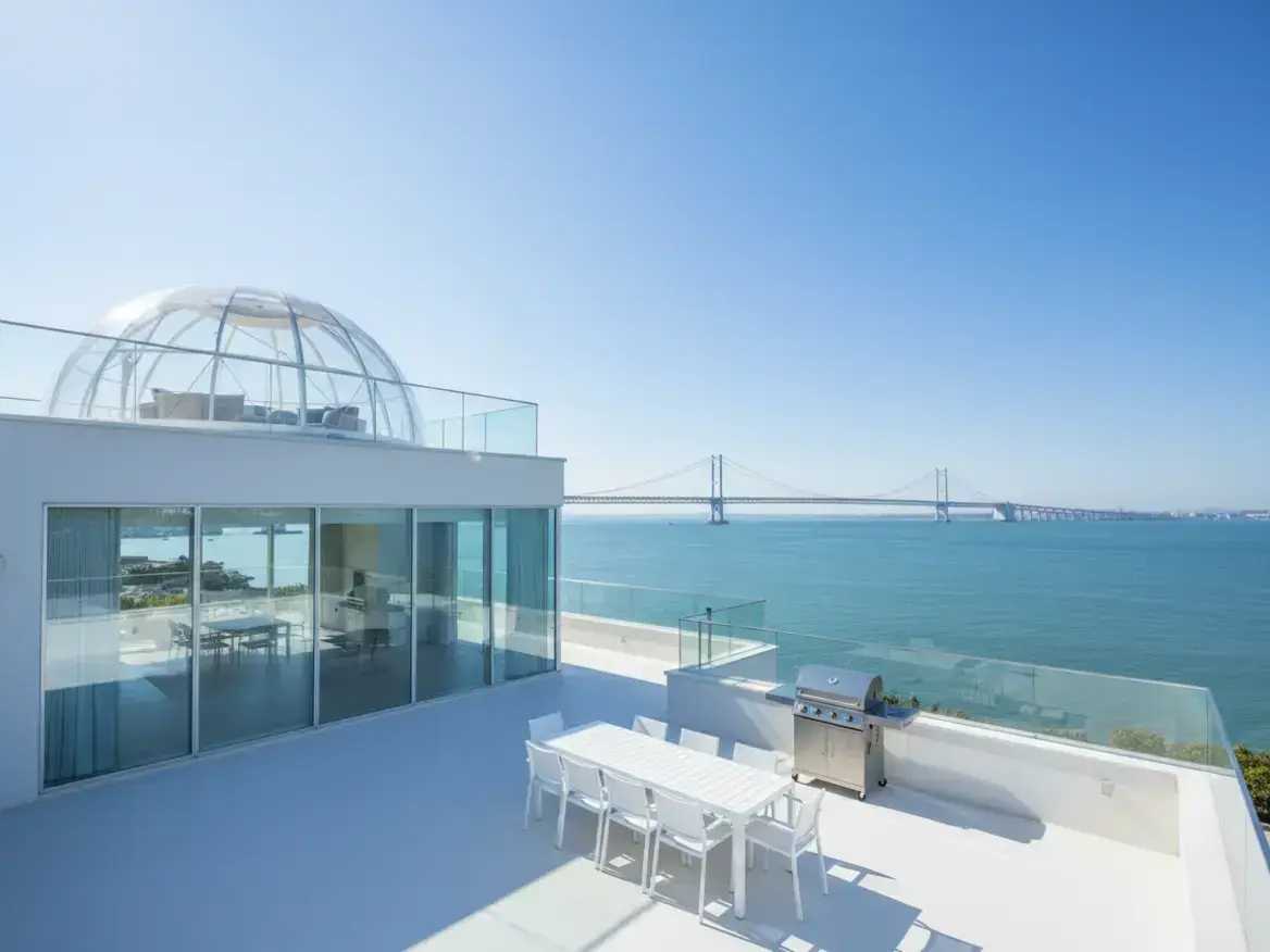 晴れた日、海に面したモダンな白い建物の屋上テラス。白いダイニングセットとバーベキューグリルが置かれ、遠くには大きな吊り橋が見える。上階にはガラスドームがある。
